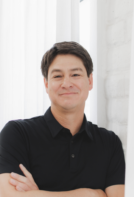 Headshot of Ray Rivera posing for a photo with his arms crossed, wearing a black shirt against a white backdrop. 