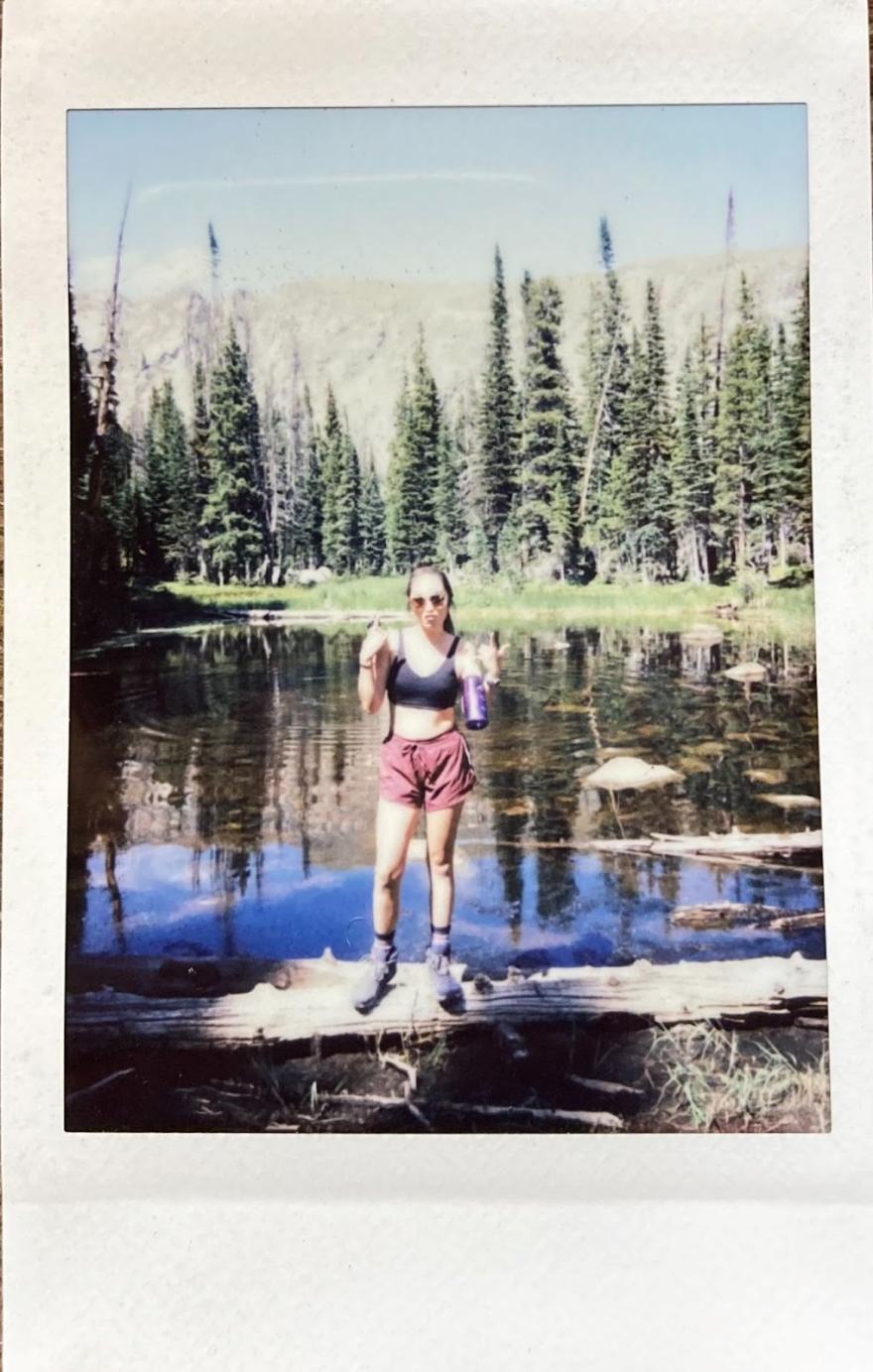 Bethany Spencer in a polaroid style photo posing at a lake with trees in the background, presumably after a hike. 