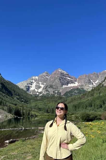 Kylie Yang wearing black sunglasses and a light yellow jacket, hair in two braids, posing in a green meadow with rocky mountains in the background during springtime. 
