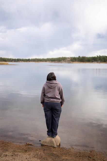 Photo taken from the back of a young woman balancing on a rock on the shore of a body of water. 